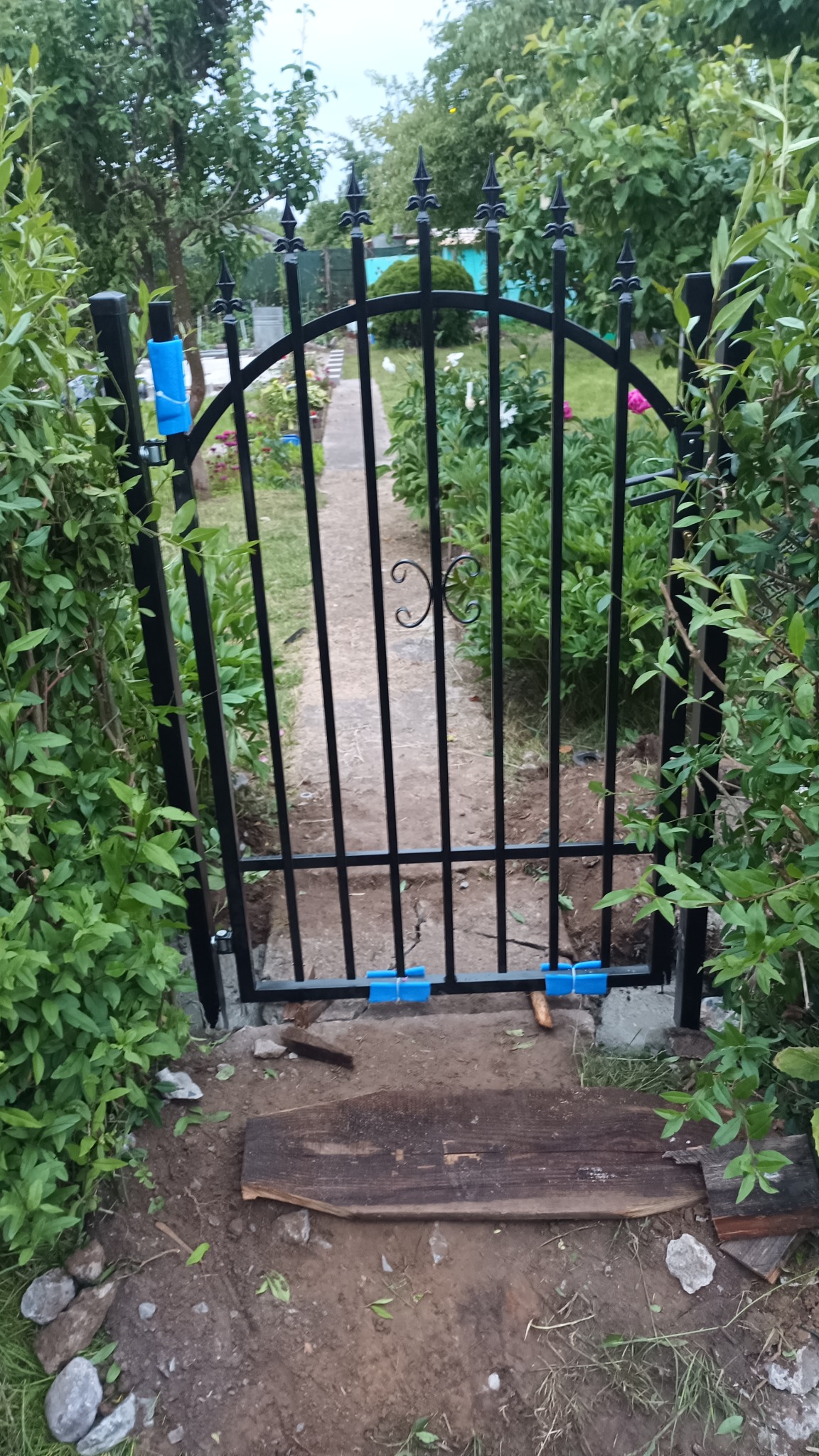 Czarna, metalowa furtka ogrodowa z ozdobnymi grotami i elementem dekoracyjnym pośrodku, zabezpieczona niebieską folią ochronną, prowadząca do ogrodu z widoczną ścieżką i kwiatami.