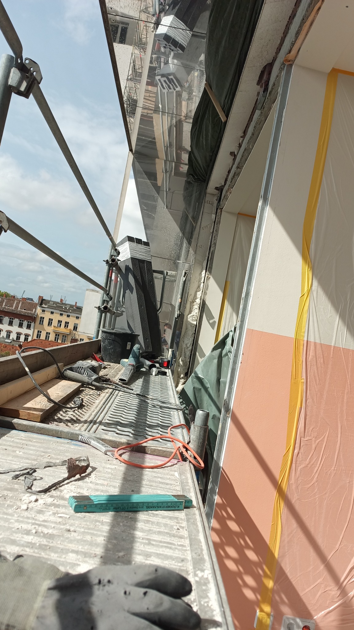 Widok z rusztowania podczas prac remontowych okna, z widocznymi narzędziami, osłoniętą folią ścianą i odbiciem budynku w oknie