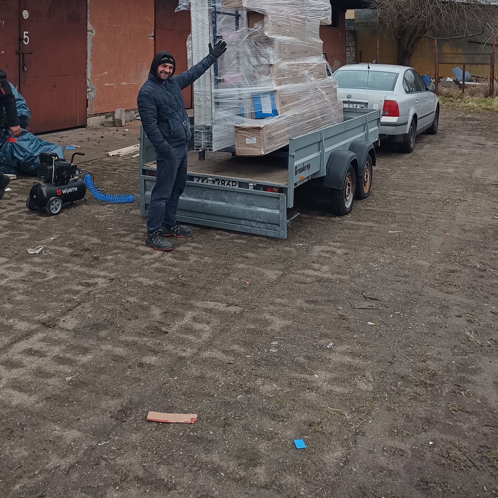 Uśmiechnięty mężczyzna w kurtce stoi obok przyczepy z załadowanymi oknami PCV, zabezpieczonymi folią stretch, na tle garaży i samochodu osobowego.
