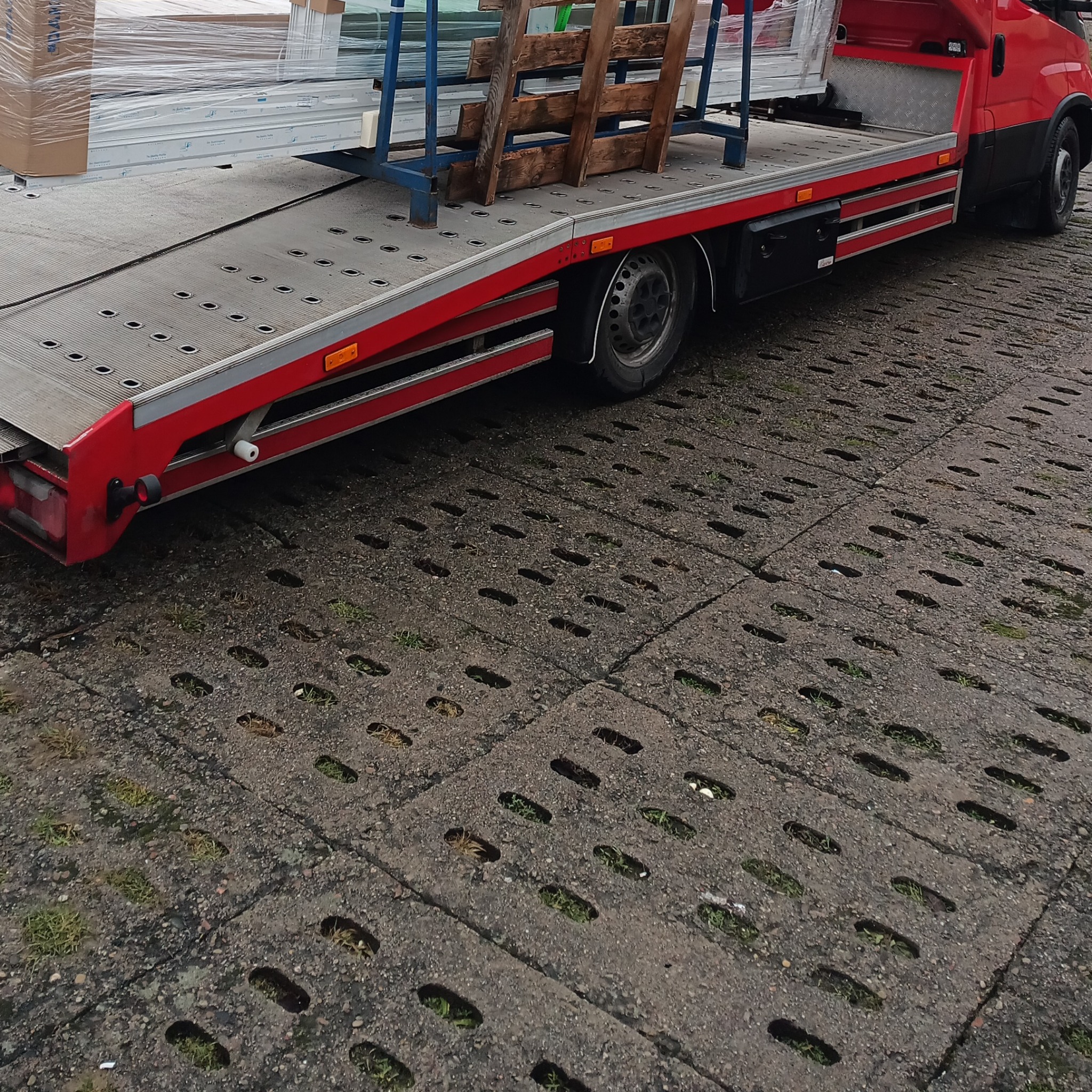 Czerwony samochód dostawczy z platformą transportową przewożący okna w opakowaniach, zabezpieczone drewnianą konstrukcją, na tle betonowej nawierzchni z otworami.