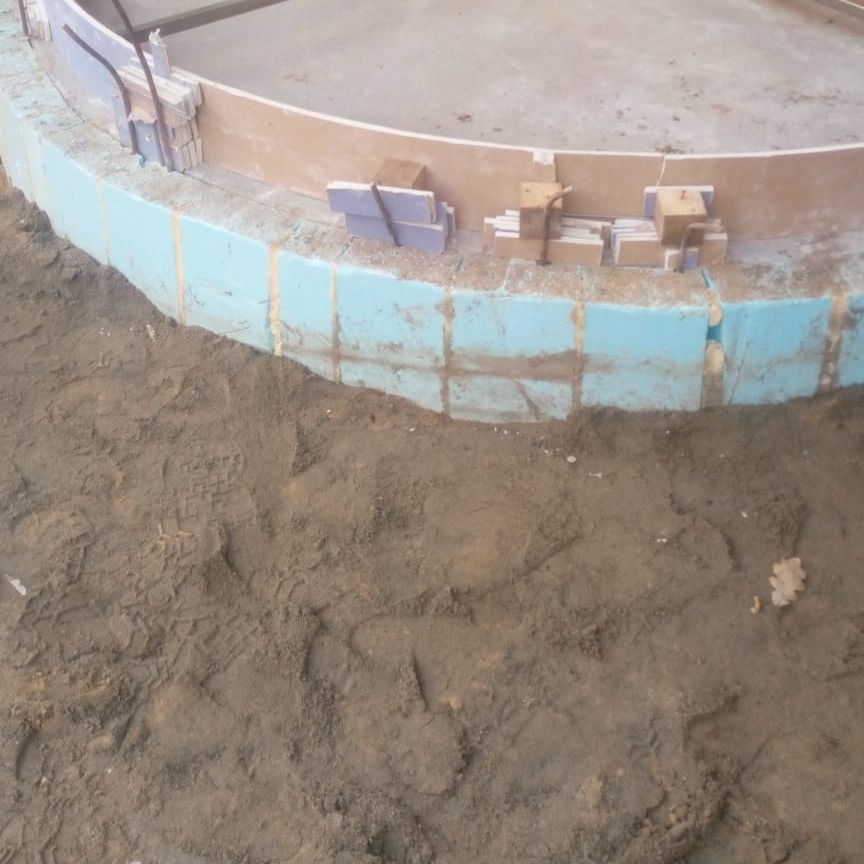 Budowa okrągłego basenu: niebieski styropianowy szalunek, betonowy wieniec i przygotowane płytki na mokrym podłożu z odciskami butów.