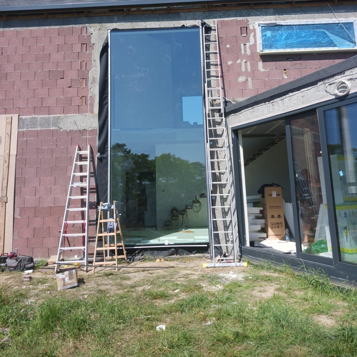 Montaż dużego okna panoramicznego w surowej bryle domu z czerwonej cegły, widoczne drabiny i narzędzia, trawa na pierwszym planie.