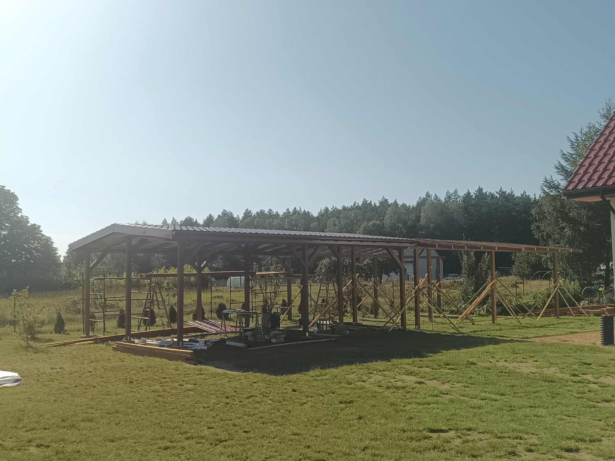 Wznoszenie drewnianej konstrukcji z zadaszeniem na trawniku, widoczne stemple budowlane, w tle las.
