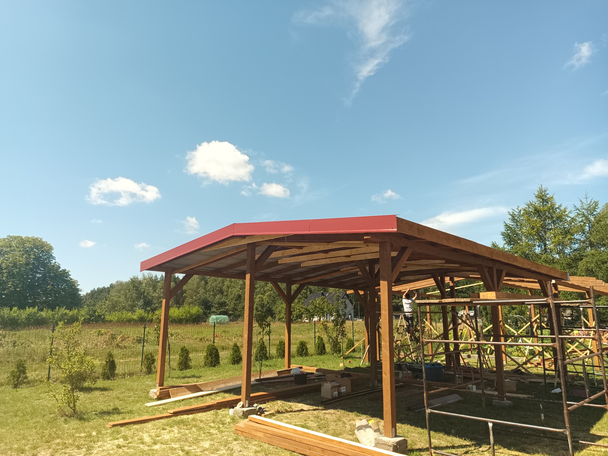 Konstrukcja drewnianej altany z czerwonym dachem w trakcie budowy, widoczna osoba na drabinie, rusztowania i materiały budowlane na trawniku, błękitne niebo z chmurami w tle.