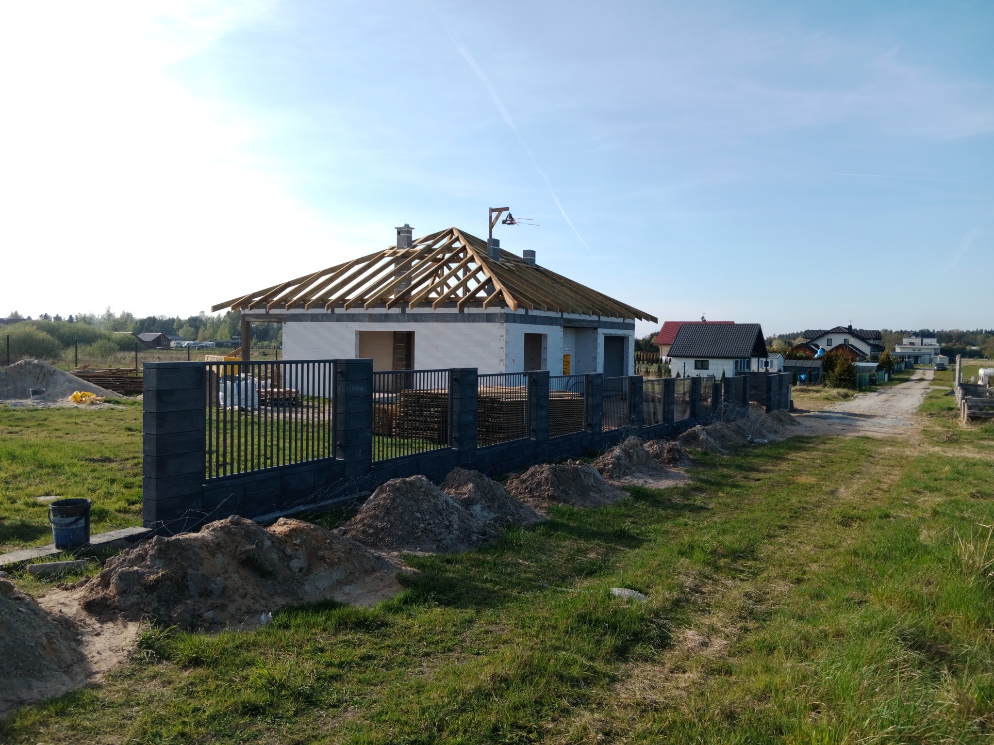 Budowa domu jednorodzinnego w trakcie realizacji: surowy stan otwarty z więźbą dachową, ogrodzenie panelowe z podmurówką, pryzmy piasku wzdłuż ogrodzenia.