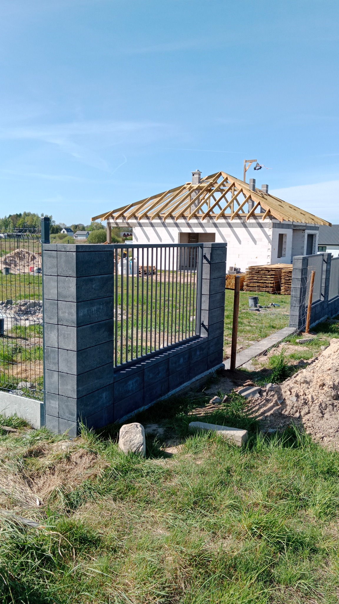 Fragment ogrodzenia panelowego z betonowymi słupkami na tle domu w trakcie budowy dachu, widok z zewnątrz posesji, trawa i ziemia na pierwszym planie.