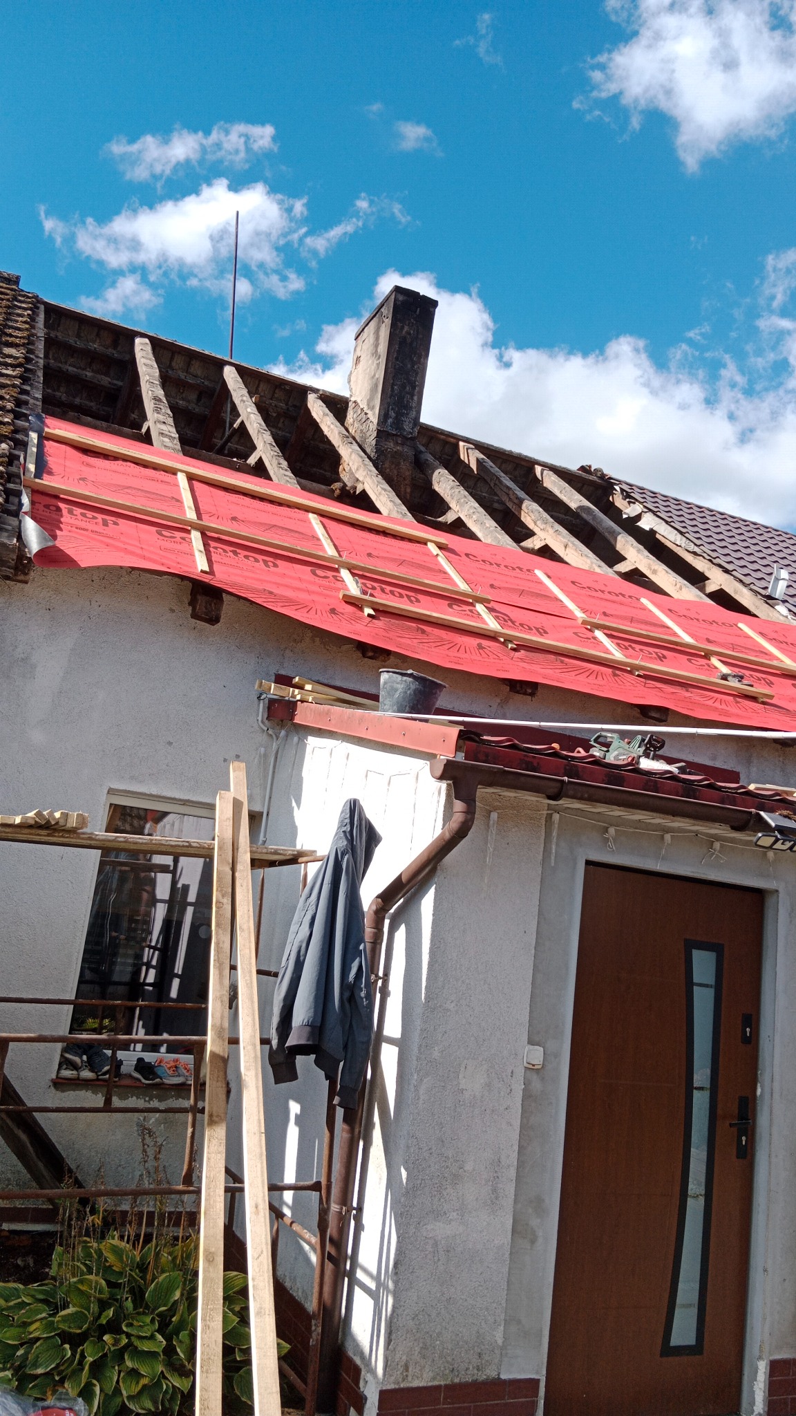 Częściowo zdjęty dach z widoczną konstrukcją i czerwoną membraną. Na ścianie budynku kurtka i drabina, a w tle błękitne niebo z chmurami.