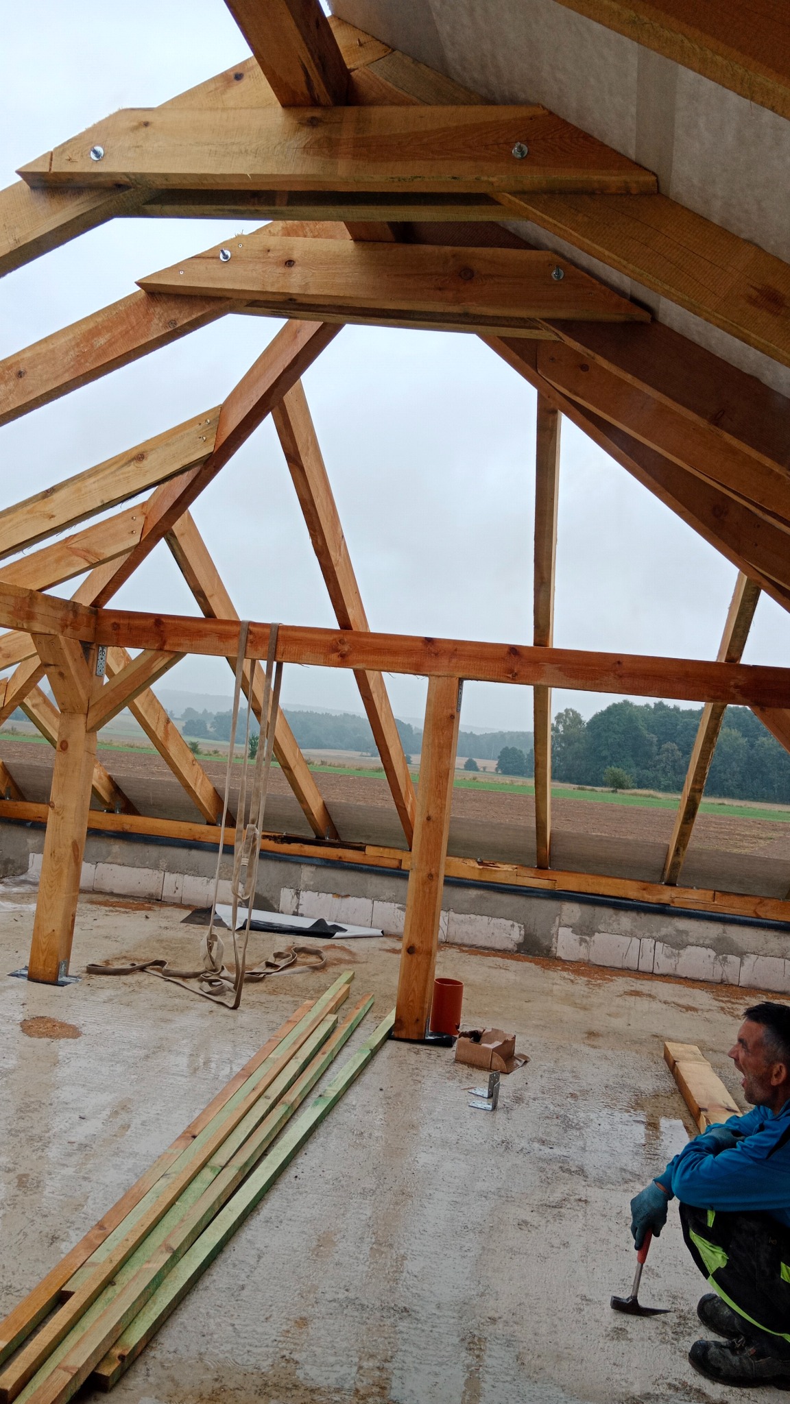 Drewniana konstrukcja dachu w trakcie budowy, widok z dołu na niebo i krajobraz. W tle pola i lasy, na pierwszym planie mężczyzna z młotkiem.