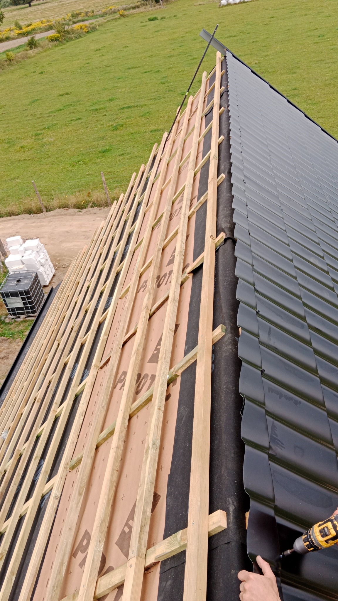 Montaż czarnej blachodachówki na drewnianej konstrukcji dachu, widok z góry na częściowo pokryty dach i zielone łąki w tle. Prace dekarskie w toku.