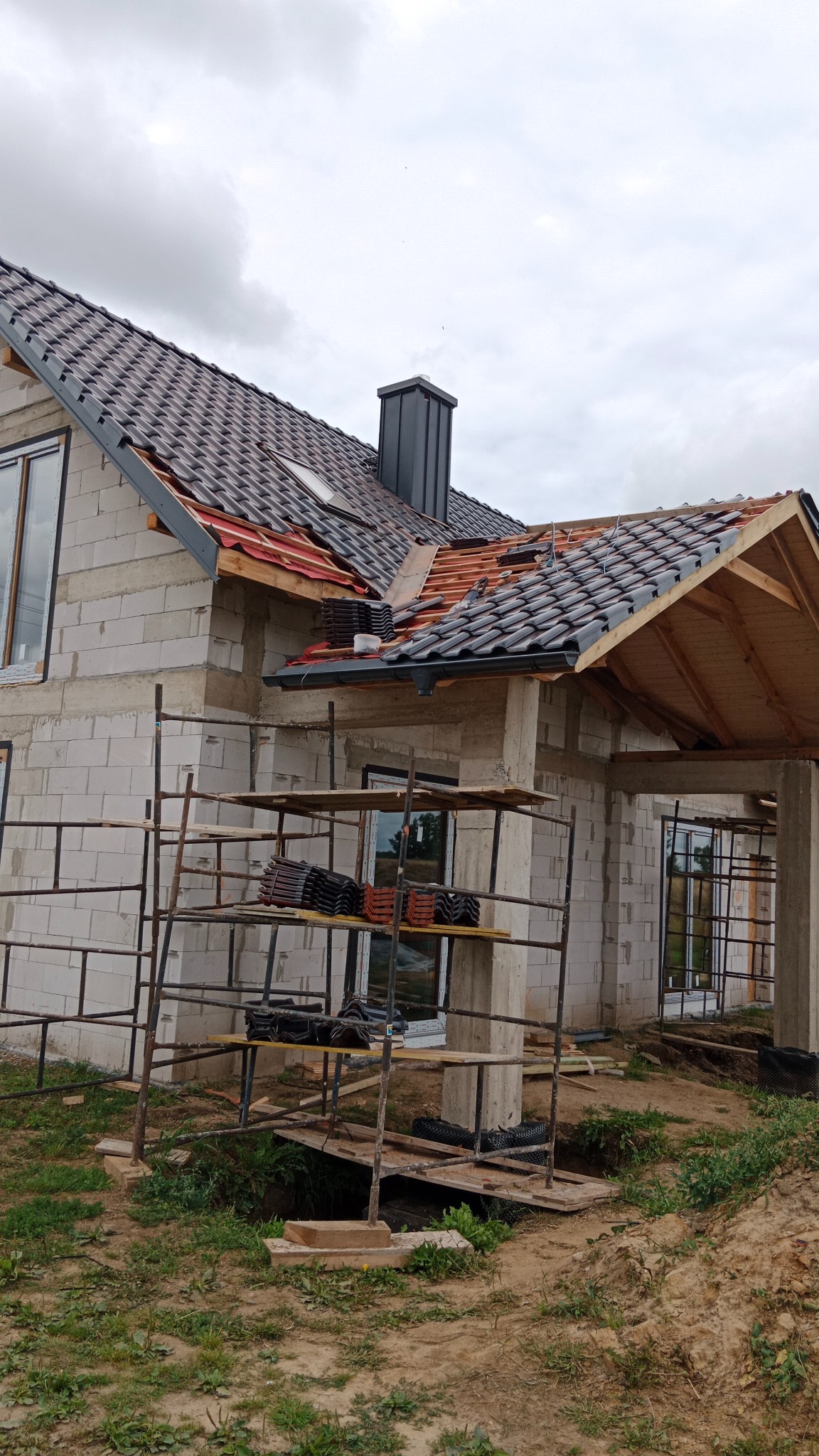 Budowa domu w trakcie krycia dachu: widoczne rusztowania, ściany z bloczków, częściowo ułożona dachówka w kolorze grafitowym i czarny komin na tle pochmurnego nieba.