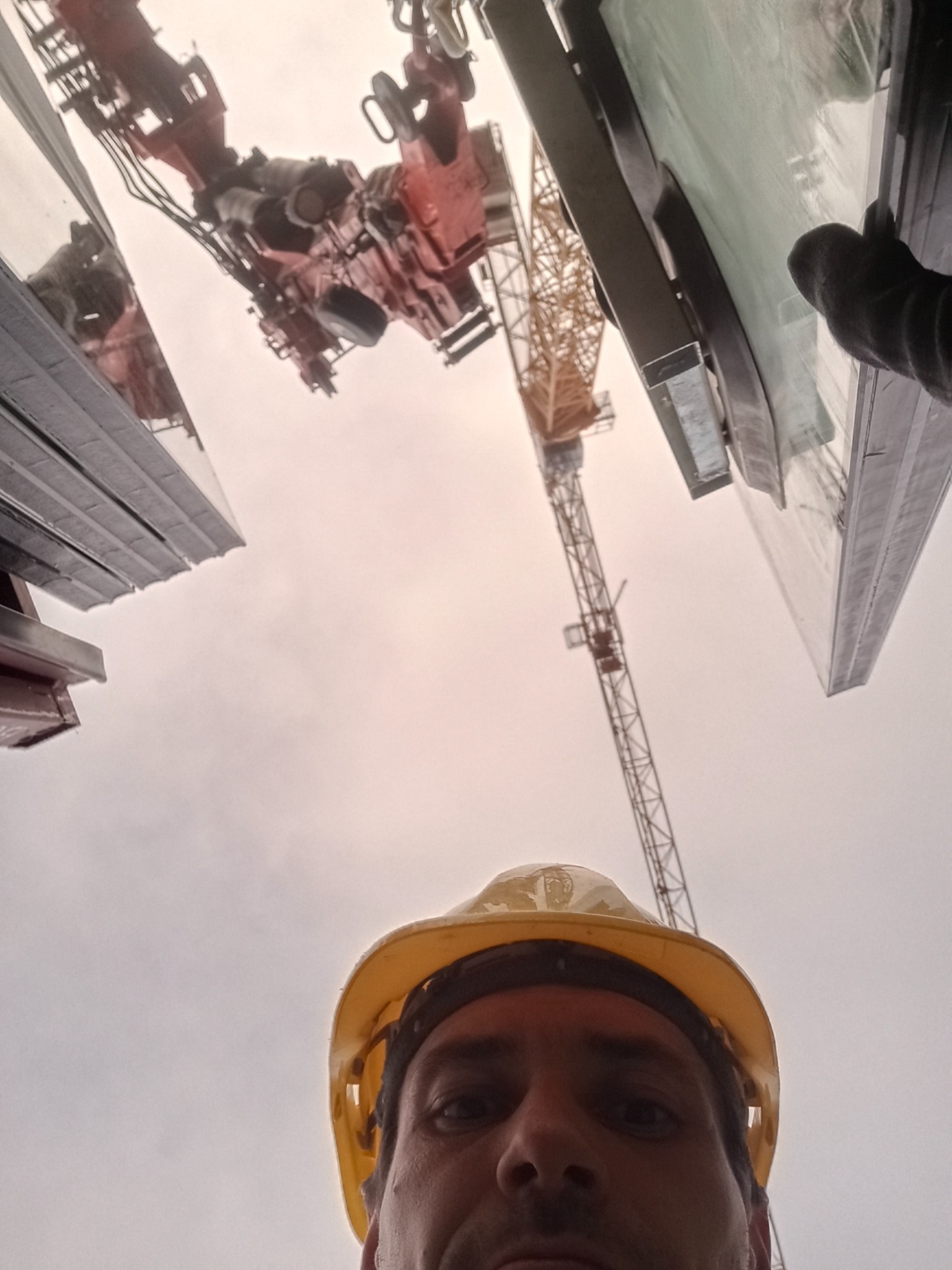 Autoportret pracownika w kasku na tle żurawia i elementów konstrukcyjnych budynku w trakcie budowy, z perspektywy żabiej.