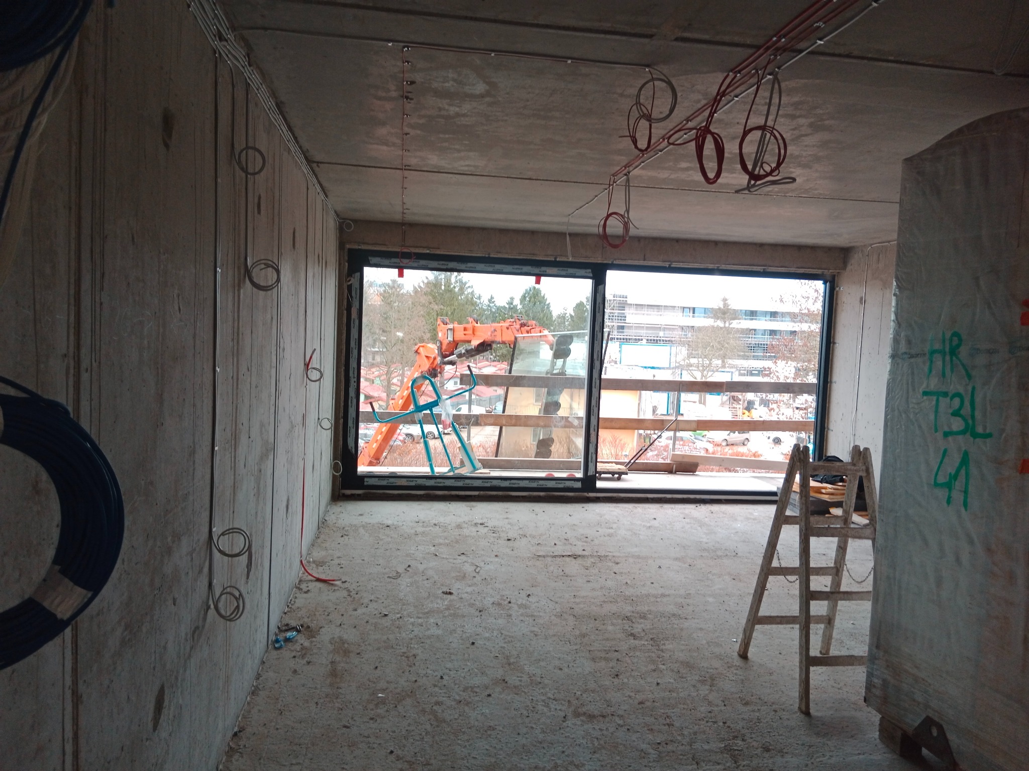 Surowy stan wnętrza z widoczną instalacją elektryczną na suficie, oknem z widokiem na dźwig i drabiną opartą o ścianę. W tle budynki.