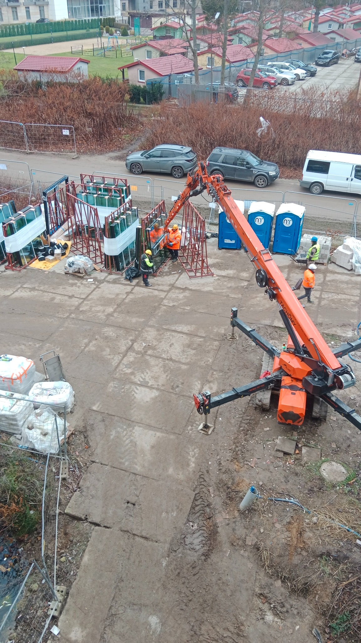 Montaż dużych przeszkleń na budowie z użyciem dźwigu. Widoczni pracownicy w kaskach i odblaskowych ubraniach. Na drugim planie budynki mieszkalne i zaparkowane samochody.