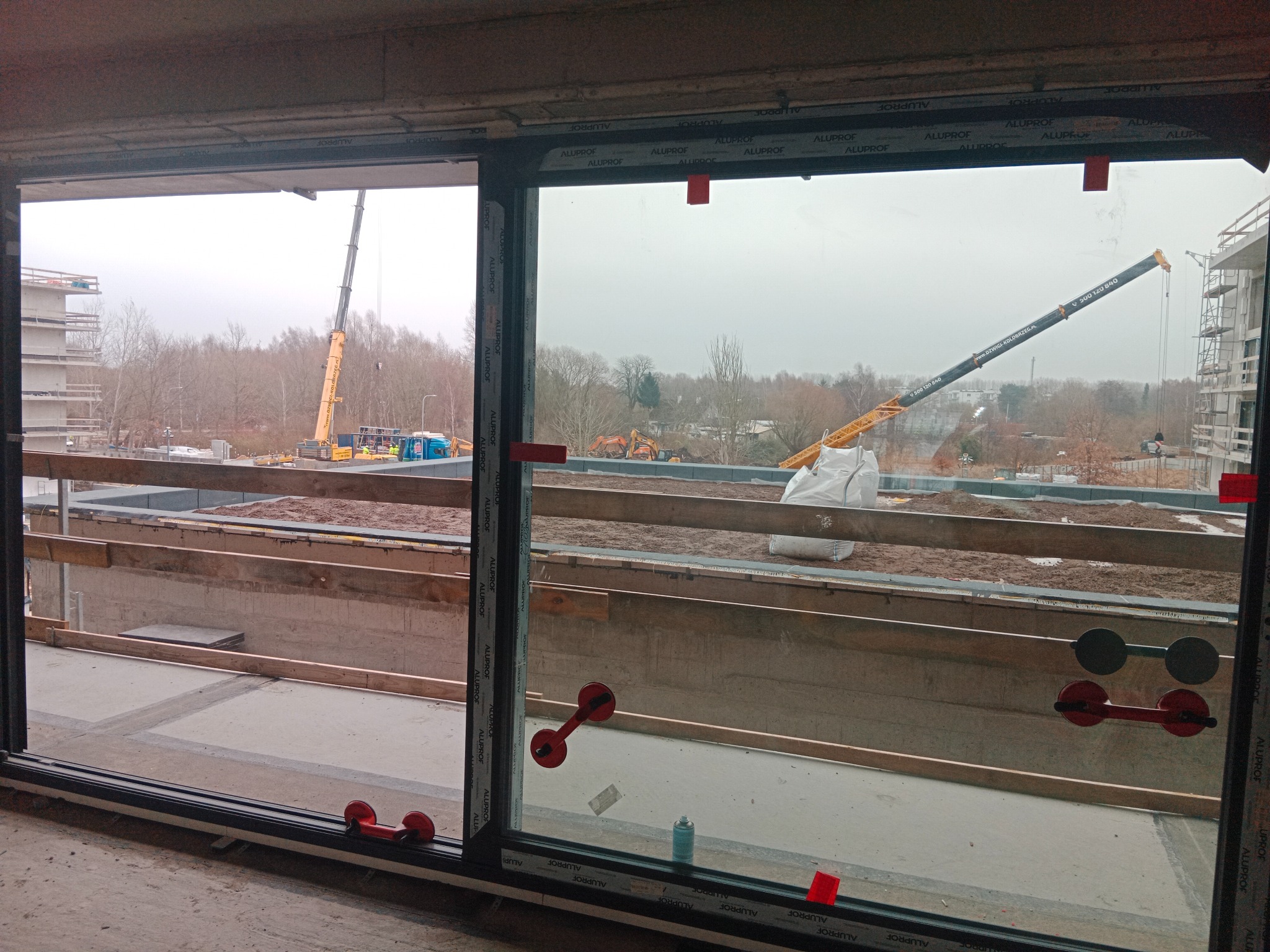 Montaż okna panoramicznego z przyssawkami w surowym wnętrzu. W tle widoczny żuraw budowlany i budowa w toku, co sugeruje etap prac budowlanych.