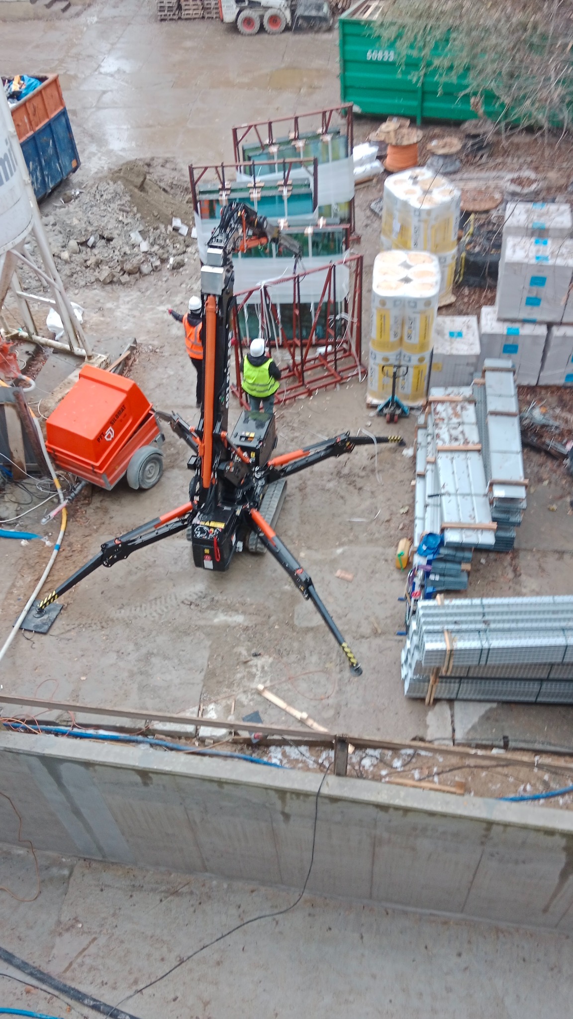 Przenośny żuraw dekarski na budowie, z pracownikami w kamizelkach odblaskowych. W tle materiały budowlane i kontenery. Widok z góry, pochmurny dzień.