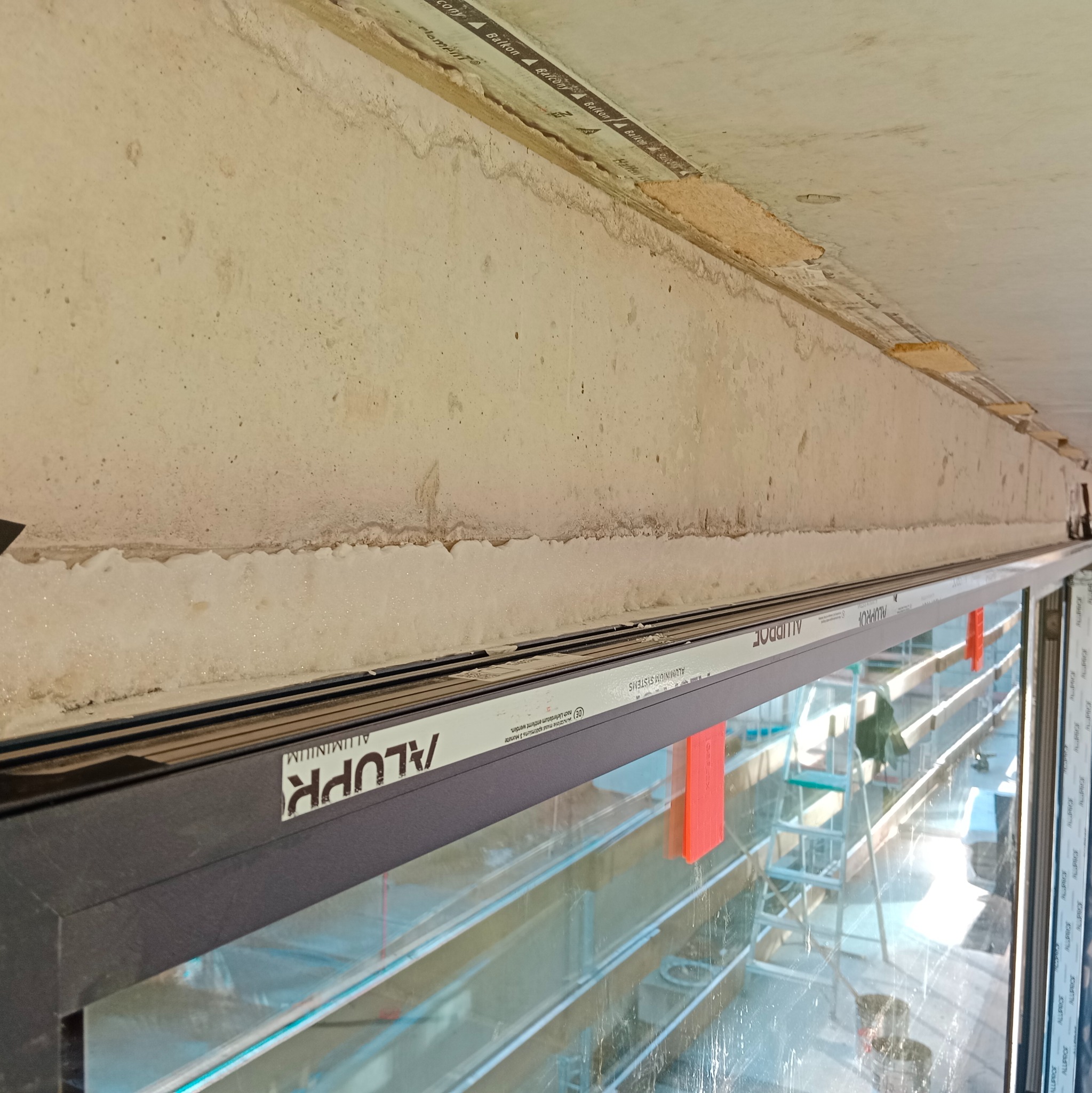 Szczegółowe ujęcie okna balkonowego z widoczną ramą aluminiową ALUPROF i fragmentem surowego sufitu. Widoczne ślady po montażu i elementy konstrukcyjne.