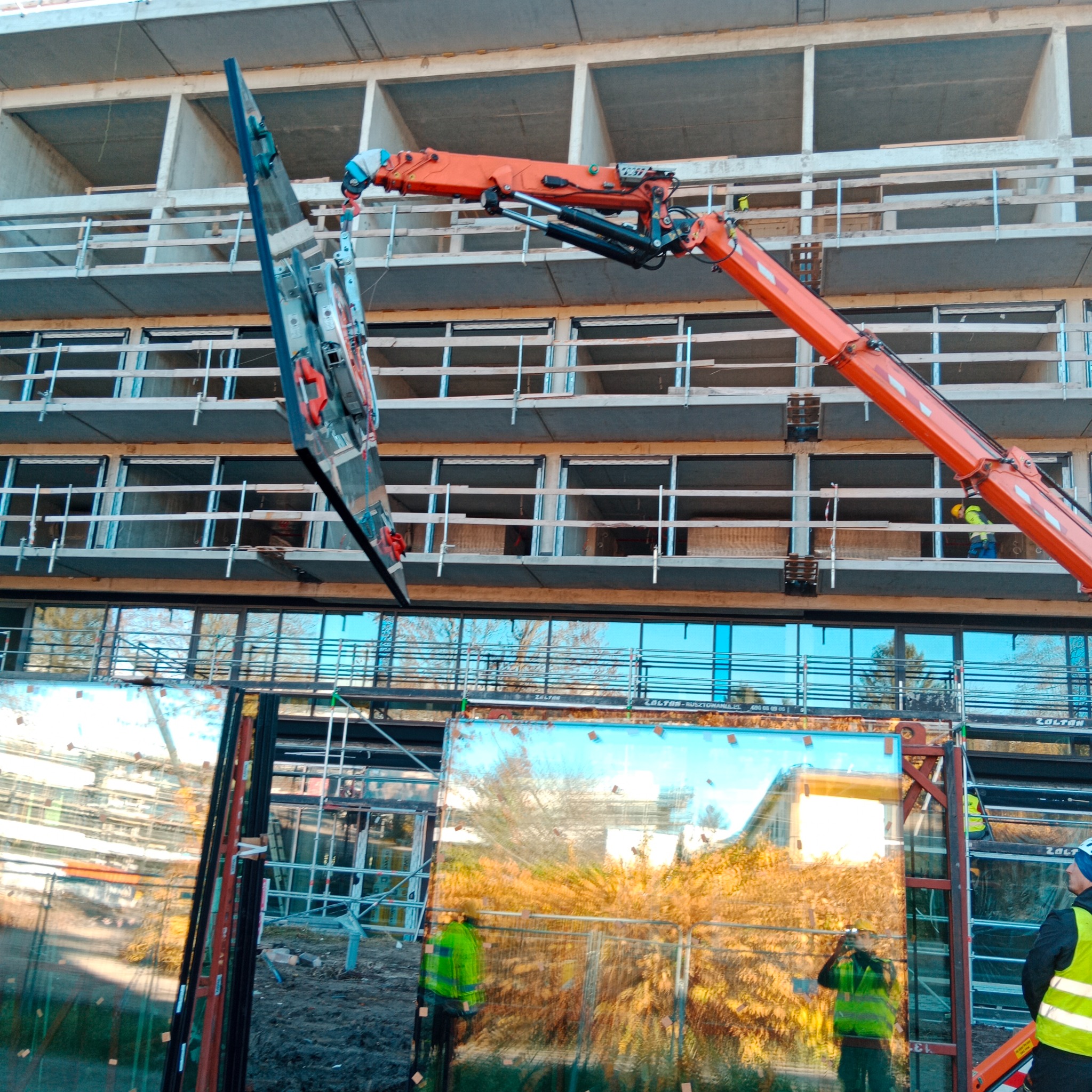 Montaż dużych szklanych paneli na fasadzie budynku za pomocą dźwigu. Widoczne rusztowania i pracownicy w odblaskowych kamizelkach. Budynek w stanie surowym.