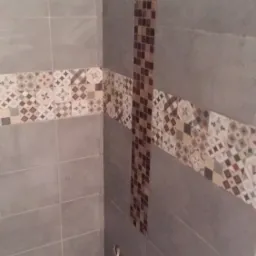 Świeżo wykafelkowana ściana łazienki z poziomym pasem dekoracyjnym i pionowym paskiem mozaiki, widoczny fragment podłogi z wzorzystymi płytkami i wystający kabel.