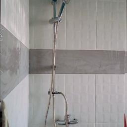 Poldom - Zamontowany panel prysznicowy z deszczownicą i słuchawką na tle białych płytek z szarym pasem dekoracyjnym, widoczny fragment niezakończonego wykończenia pod panelem i poziomica w rogu.