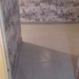 Fragment pokoju z tapetą w szaro-białe wzory, szarą podłogą z płytek i małym lustrem w drewnianej ramie na ścianie.