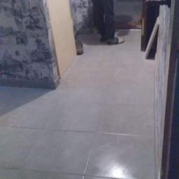 Poldom - Świeżo położone, błyszczące, szare płytki podłogowe w łazience, widoczna osoba w czerwonej bluzce pochylona nad podłogą, prawdopodobnie podczas prac wykończeniowych.