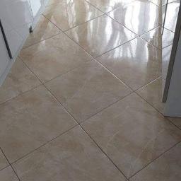 Poldom - Błyszcząca, beżowa podłoga z płytek ułożonych w karo, widok z perspektywy podłogi, z fragmentem białych szafek dolnych kuchennych i suszarki na pranie w tle.