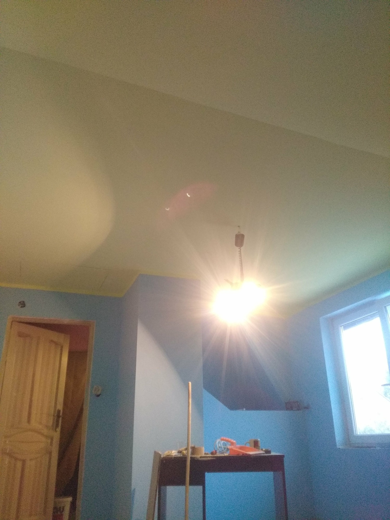 Pokój w trakcie odświeżania: ściany pomalowane na niebiesko, z żółtą taśmą malarską przy suficie; widoczny stół z narzędziami i otwarte drewniane drzwi.