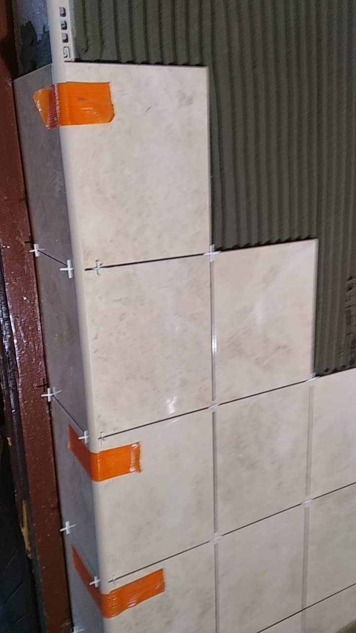 Układanie beżowych płytek w łazience, widoczne krzyżyki dystansowe i pomarańczowa taśma zabezpieczająca narożniki, świeżo nałożony klej na ścianie.