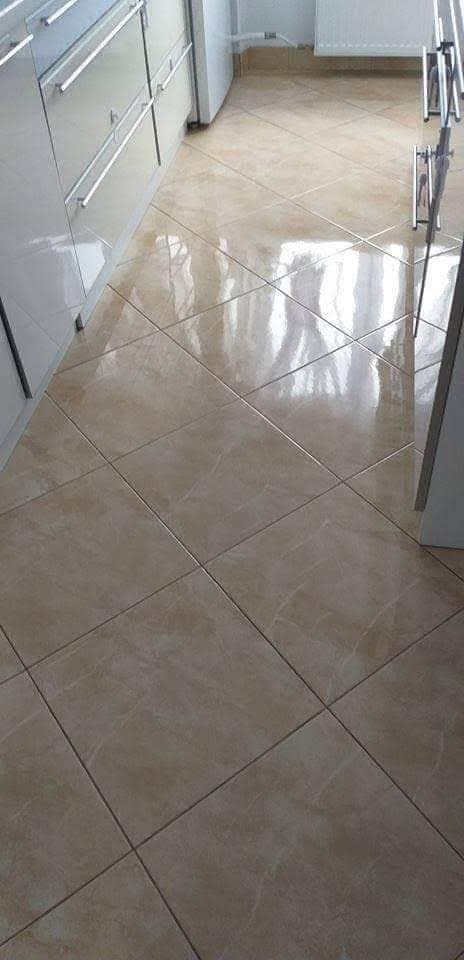 Błyszcząca, beżowa podłoga z płytek ułożonych w karo, widok z perspektywy podłogi, z fragmentem białych szafek dolnych kuchennych i suszarki na pranie w tle.