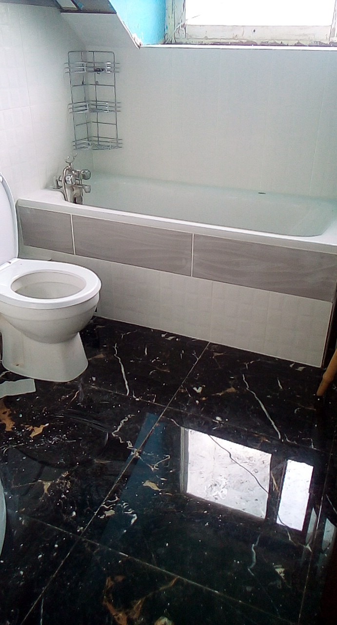 Biała toaleta z otwartą klapą obok wanny z szarymi panelami, na podłodze ciemne płytki imitujące marmur.