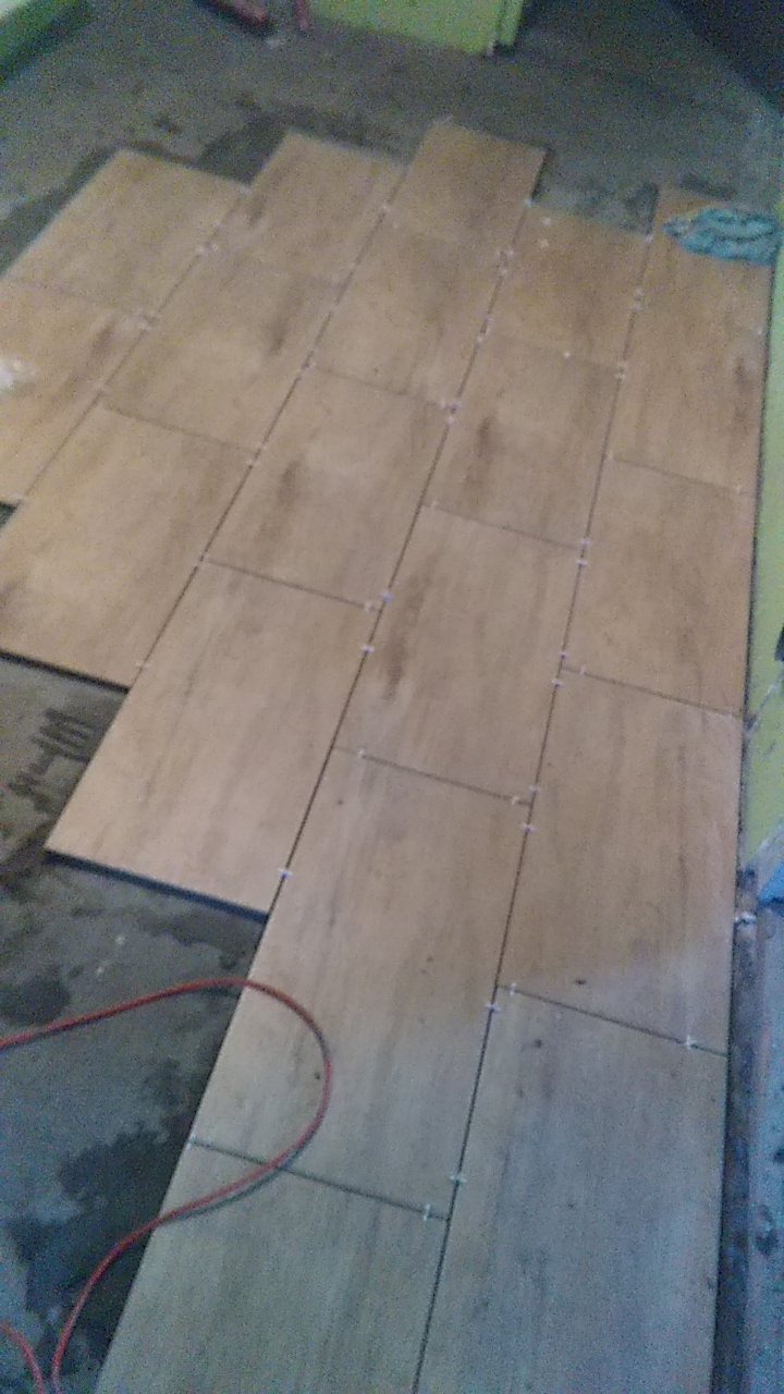 Układanie podłogowych płytek imitujących drewno z krzyżykami dystansowymi w trakcie prac wykończeniowych, widoczny fragment czerwonego kabla i mokra podłoga.