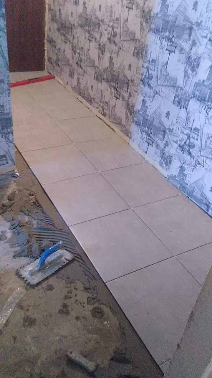 Układanie jasnych, kwadratowych płytek podłogowych w wąskim pomieszczeniu, widoczna szpachelka i zaprawa, ściana w tle pokryta tapetą z motywem miejskim.