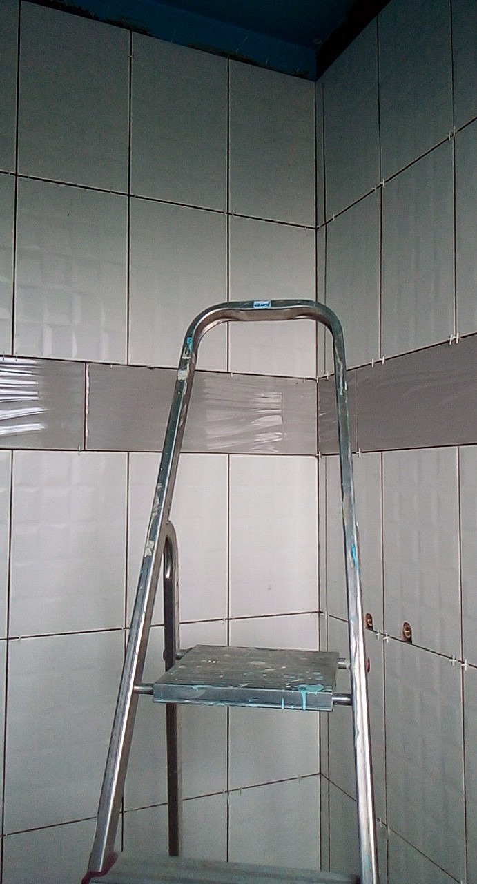 Świeżo ułożone, białe, błyszczące płytki w łazience z szarą dekoracyjną listwą, widoczne krzyżyki dystansowe i metalowa drabina z plamami zaschniętego kleju.