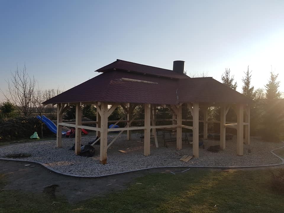 Drewniana konstrukcja altany z dachem pokrytym czerwoną dachówką bitumiczną w ogrodzie, na podłożu wysypanym drobnym żwirem, w tle widoczna zjeżdżalnia dla dzieci.