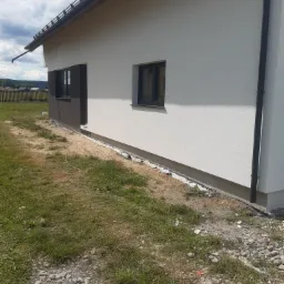 Fragment elewacji domu po tynkowaniu z widoczną listwą startową i fragmentem instalacji odprowadzającej wodę, na tle trawnika i nieba.
