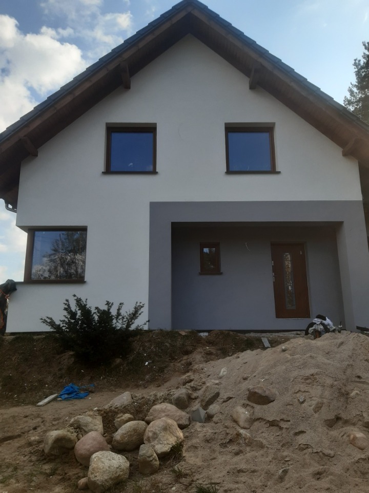 Dwukondygnacyjny dom z jasną elewacją i ciemnoszarą wnęką wejściową, widoczny fragment dachu i okien, na pierwszym planie hałda piasku i kamienie.