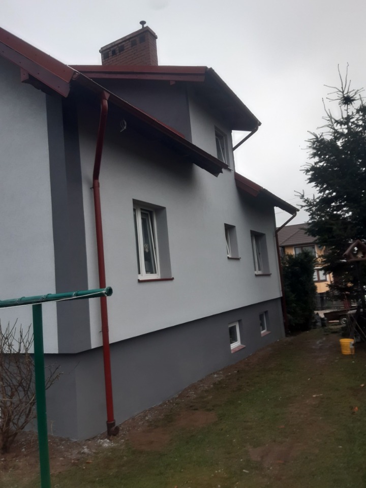 Dwukolorowa elewacja domu jednorodzinnego w odcieniach szarości, z widocznym kominem i elementami orynnowania w kolorze ceglastym, sfotografowana w pochmurny dzień.