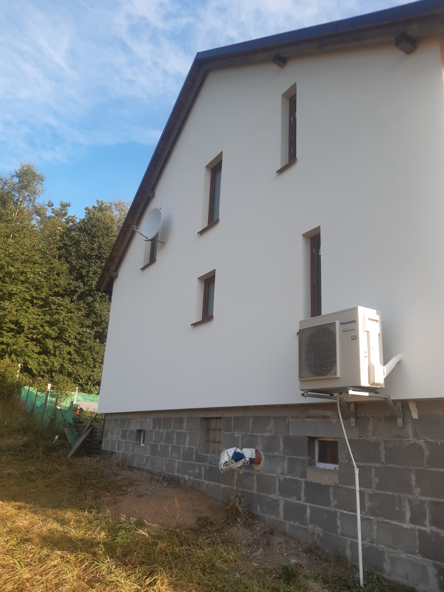 Boczny widok domu z białą elewacją, małymi, wąskimi oknami z brązowymi ramami, anteną satelitarną i zamontowaną jednostką zewnętrzną klimatyzacji Panasonic, posadowionego na fundamencie z szarych...