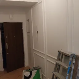 Wykończone wnętrze mieszkania z drewnianą podłogą ułożoną w jodełkę, białe ściany z dekoracyjnymi listwami, drabina oparta o ścianę i torba budowlana na podłodze.