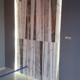 Ściana wyłożona płytkami imitującymi marmur z podświetleniem LED po bokach, widoczne elementy instalacji elektrycznej, fragment podłogi wyłożonej płytkami oraz zabezpieczenie z niebieskiej taśmy.