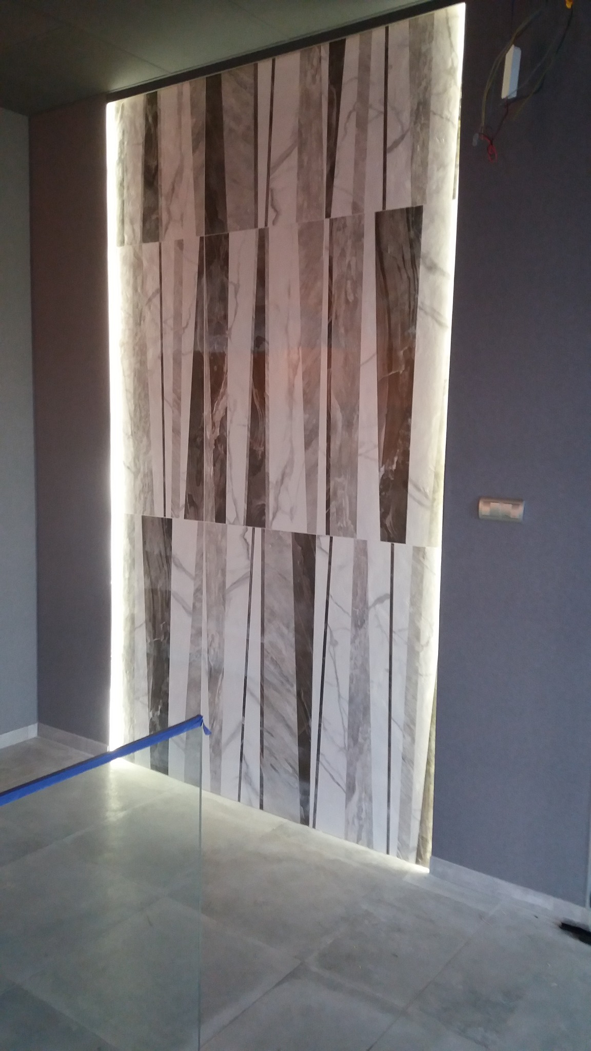 Ściana wyłożona płytkami imitującymi marmur z podświetleniem LED po bokach, widoczne elementy instalacji elektrycznej, fragment podłogi wyłożonej płytkami oraz zabezpieczenie z niebieskiej taśmy.