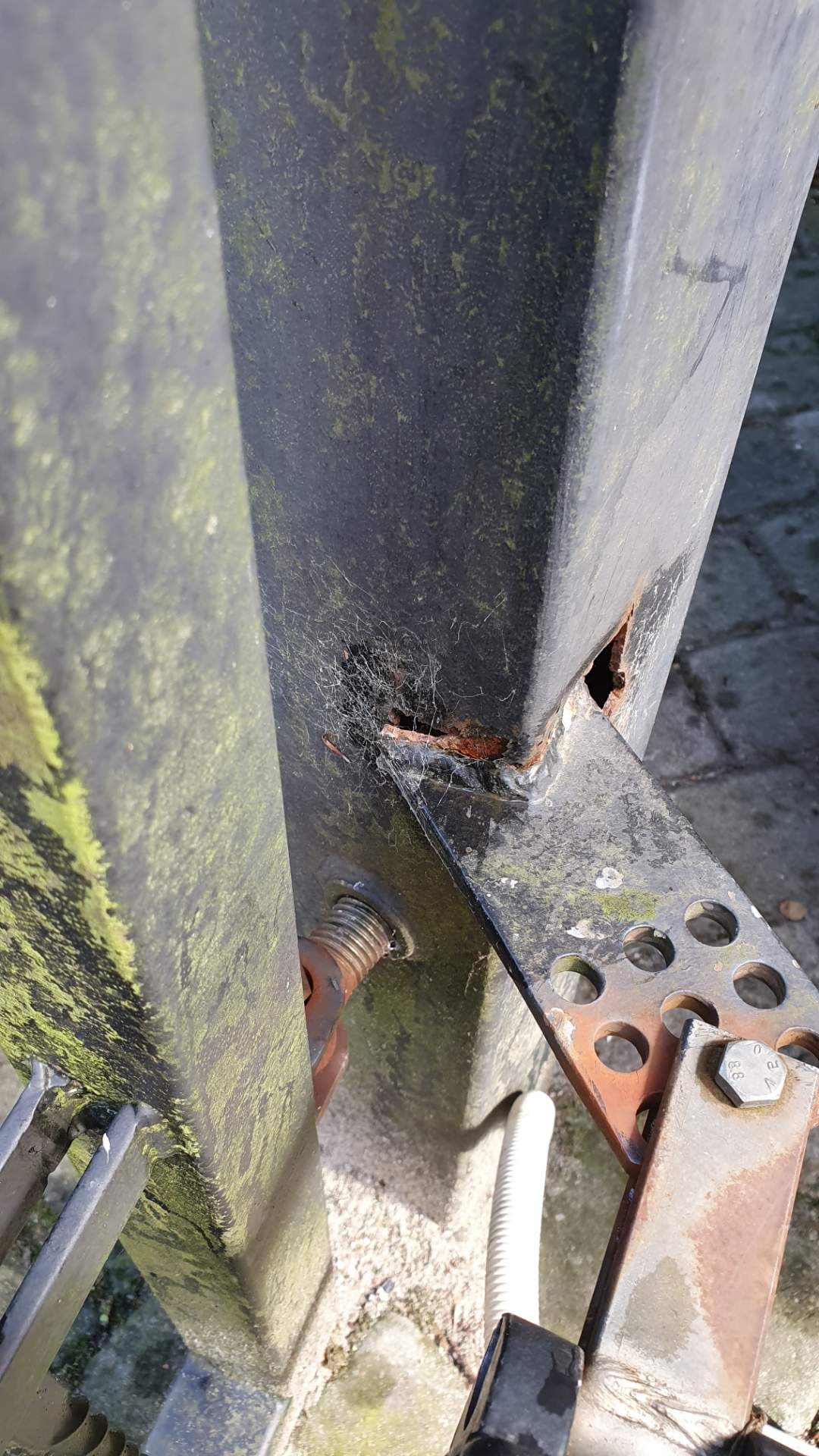 Zardzewiały, spawany element konstrukcyjny bramy z ubytkiem metalu, pokryty mchem i pajęczynami, widoczny fragment zardzewiałej śruby mocującej.