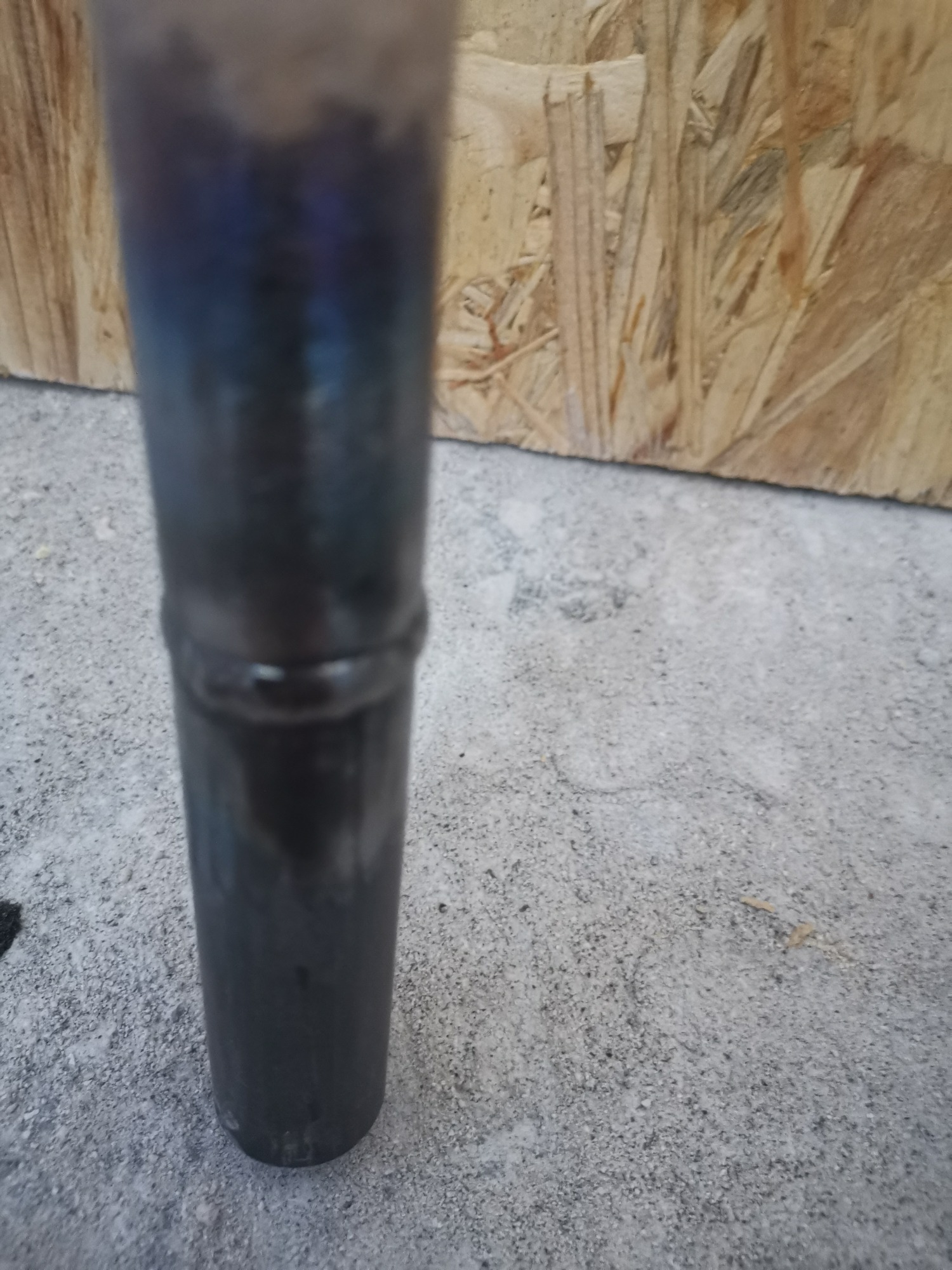 Metalowa rura z widocznym spoiwem spawalniczym, stojąca na betonowej powierzchni, z płytą OSB w tle.