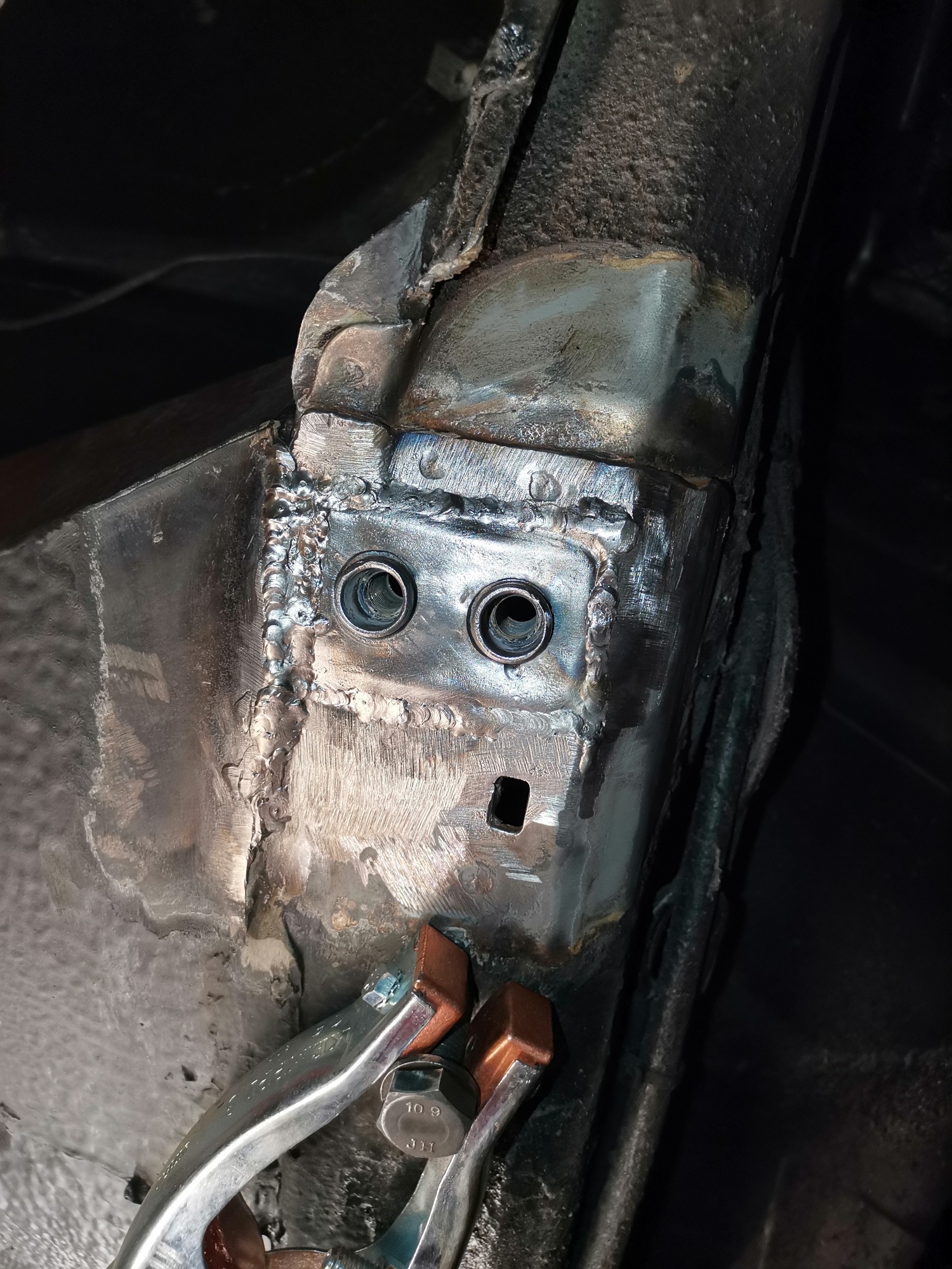 Fragment podwozia samochodu po naprawie blacharskiej, widoczne spawy wokół dwóch gwintowanych otworów, zacisk stolarski z nakrętką 10.9, surowe wykończenie metalu.
