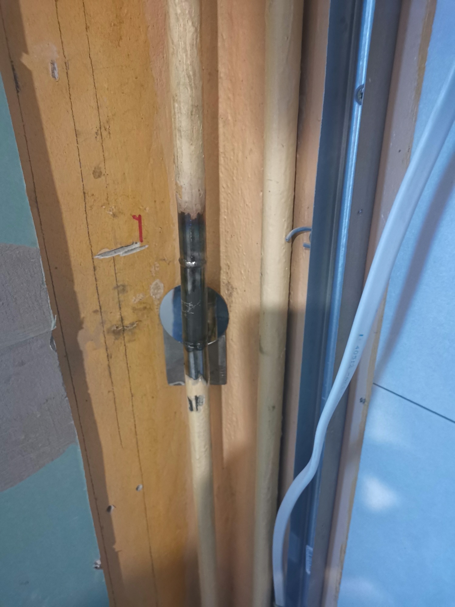 Rura połączona spawem, zamocowana do surowej ściany z widocznymi śladami po wierceniu i czerwoną naklejką, obok metalowej ramy i kabla elektrycznego.