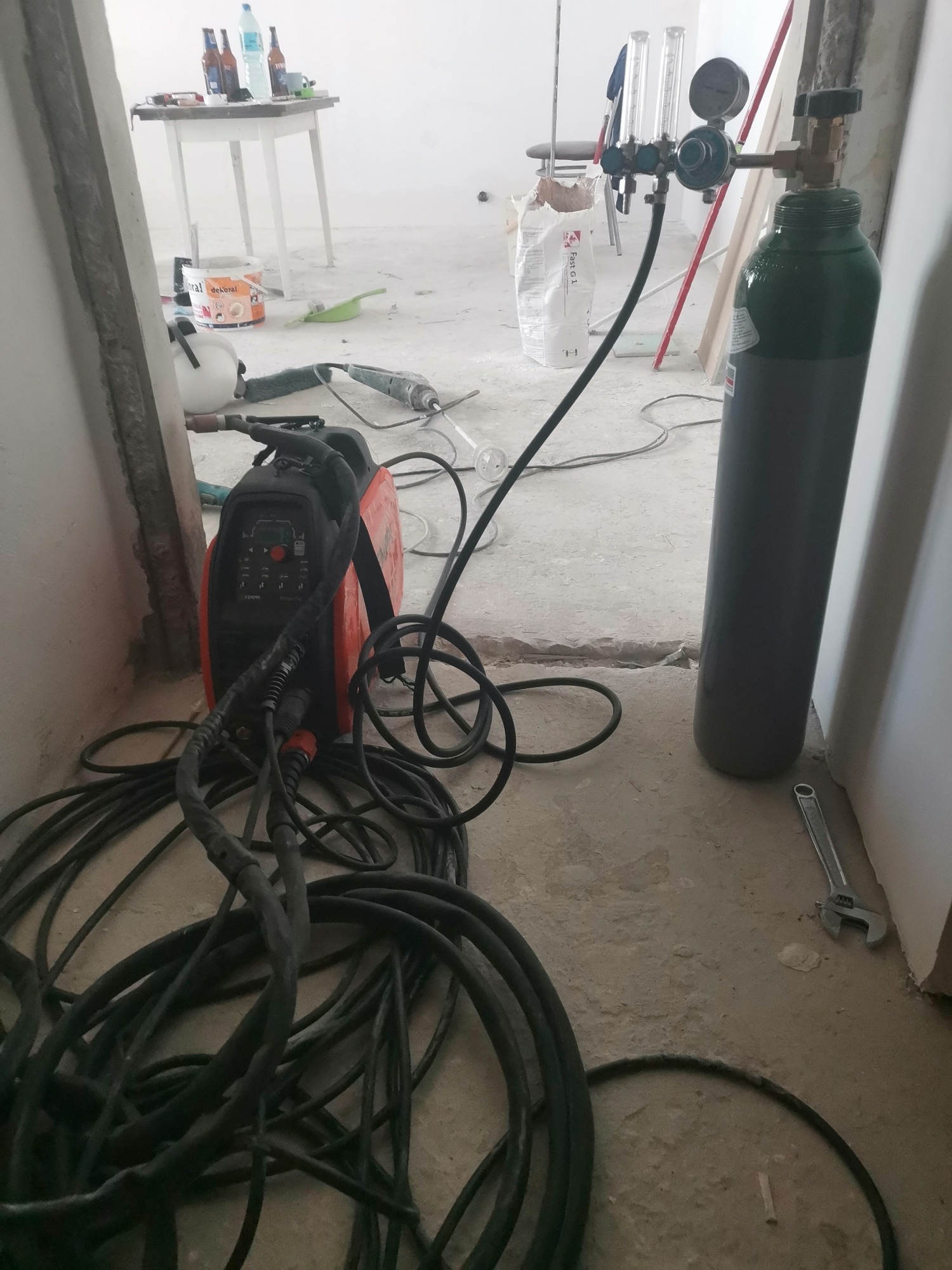 Pomieszczenie w trakcie remontu: butla z gazem technicznym, spawarka inwertorowa z przewodami, klucz nastawny leżący na podłodze.