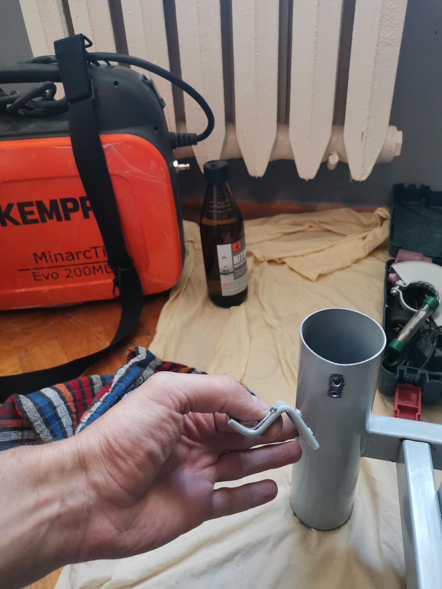 Przygotowanie do spawania metalowej konstrukcji: dłoń trzyma element mocujący przy rurze, w tle spawarka Kemppi MinarcTig Evo 200ML i grzejnik żeberkowy.