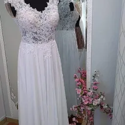 Elegancka suknia ślubna z koronkową górą i zwiewnym dołem, prezentowana na manekinie w salonie, z odbiciem innych sukien w lustrze i wazonem z różowymi kwiatami w tle.