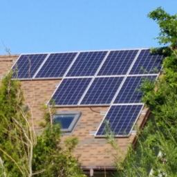 Hymon Fotowoltaika Sp. z o.o. - Panele słoneczne na brązowym dachu domu jednorodzinnego z oknem dachowym, częściowo zasłonięte przez drzewa, w słoneczny dzień.