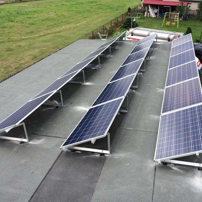 Instalacja fotowoltaiczna na płaskim dachu pokrytym papą, widok z góry, panele ustawione w trzech rzędach, w tle zielony trawnik i dom jednorodzinny.